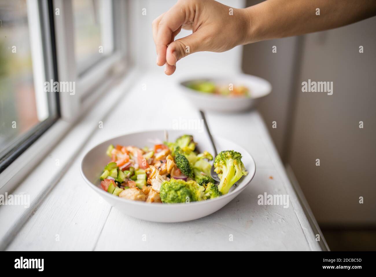 Main ajoutant une pincée de sel à un brocoli et salade de poulet à côté d'une fenêtre Banque D'Images