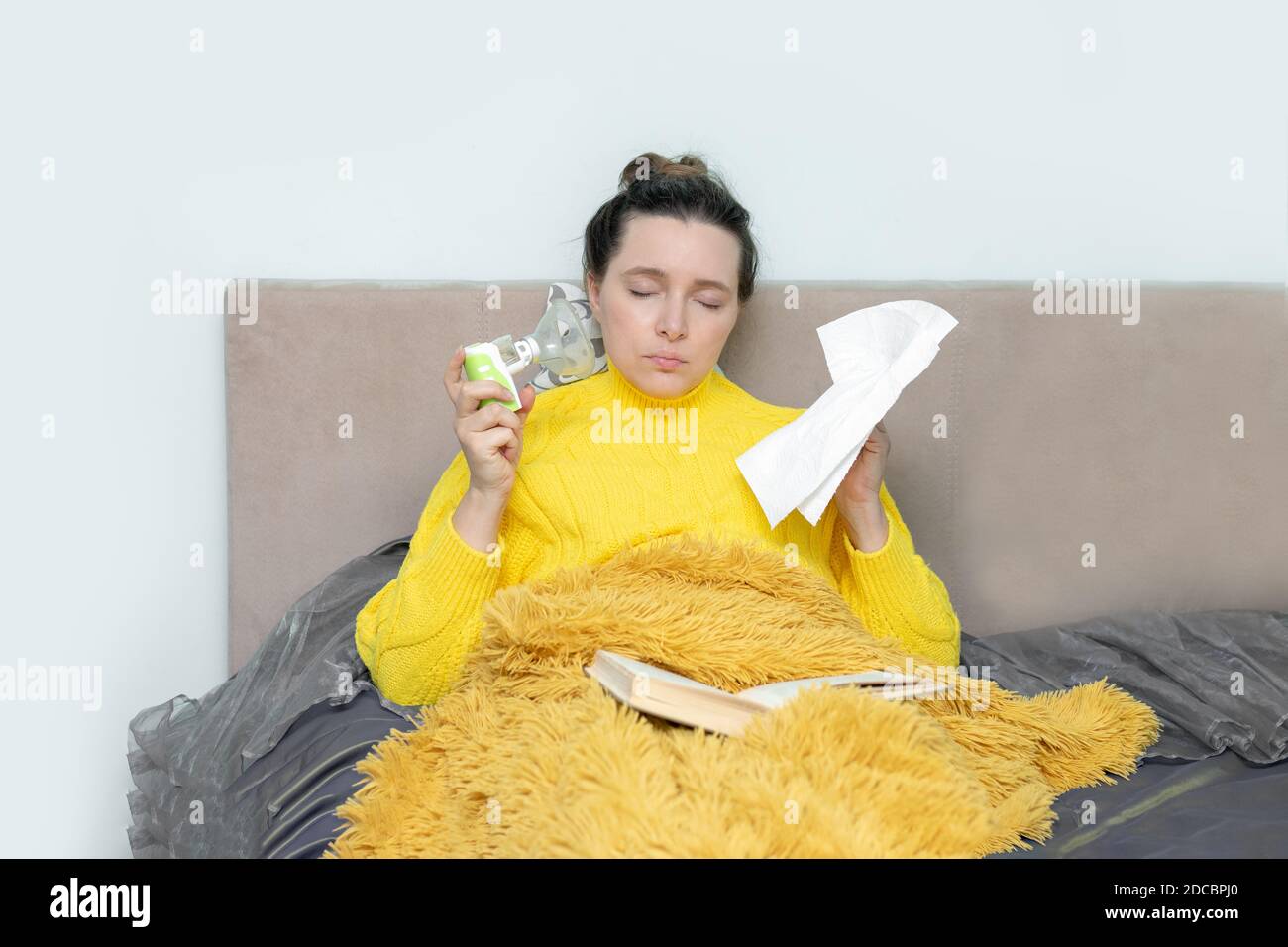 Une femme fatiguée et malade sur un lit dans un chandail et sous une couverture dans sa maison lisant un livre, une serviette en papier et un nubélizer sur l'auto-isolation pendant Banque D'Images