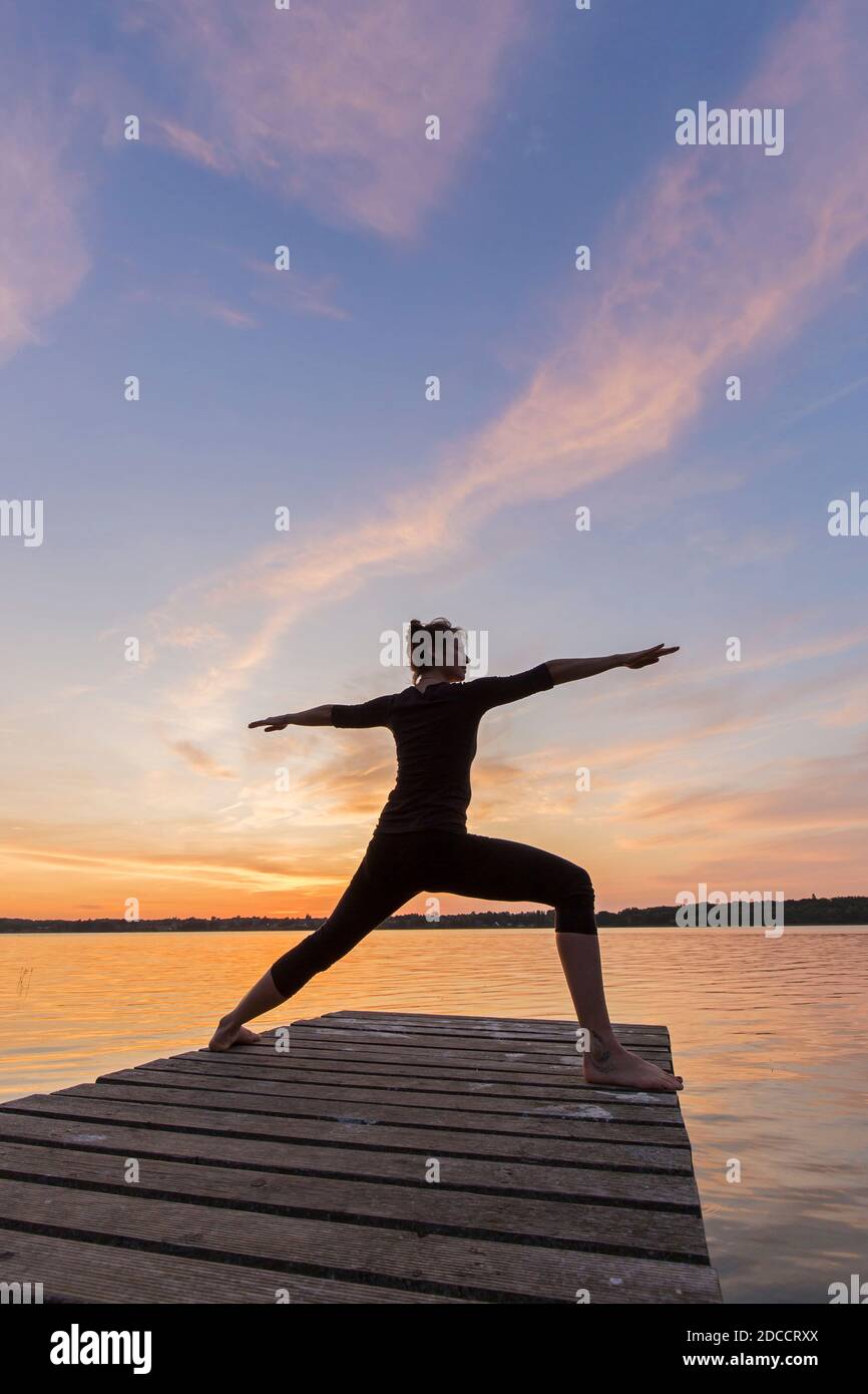 Femme pratiquant la posture de yoga Virabhadrasana II / posture du guerrier, en train de faire une fente debout à l'asana sur la jetée au lac au coucher du soleil Banque D'Images