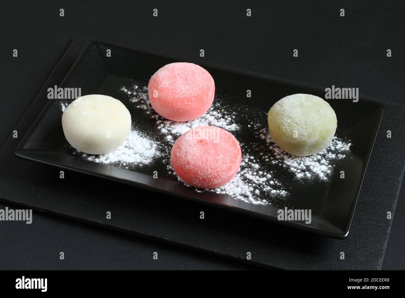 Quatre types de mochi japonais dessert - grenade au miel, thé vert matcha, fraise, noix de coco sur une assiette noire sur une table noire. Gros plan Banque D'Images