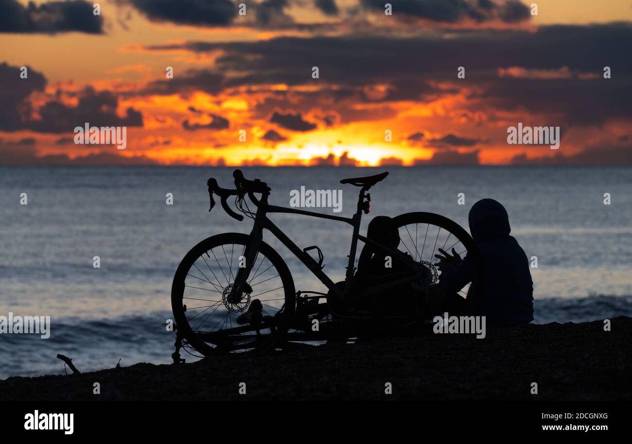 Silhouette d'une paire de cyclistes masculins reposant sur une plage tout en regardant le soleil se coucher. Les gens regardent le coucher du soleil sur la mer. Banque D'Images