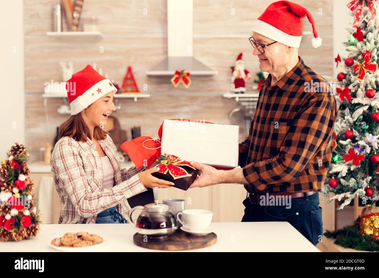 Petit-enfant heureux échangeant des cadeaux avec grand-père célébrant noël. Homme senior portant chapeau de père noël surprenant petite-fille avec des cadeaux de vacances d'hiver dans la cuisine maison avec arbre de Noël en arrière-plan. Banque D'Images