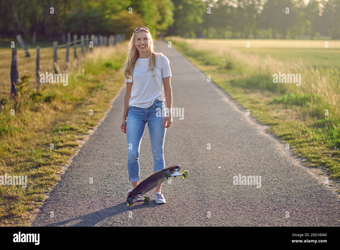 Belle femme blonde sportive avec son skateboard rétroéclairé par le le soleil du soir sur une route rurale entre les champs souriant à la caméra Banque D'Images