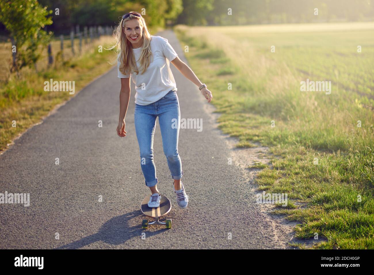FIT active femme d'âge moyen jouant sur son skateboard à l'approche de la camera le long d'une route rurale étroite avec un sourire heureux rétroéclairé par le soleil du soir Banque D'Images