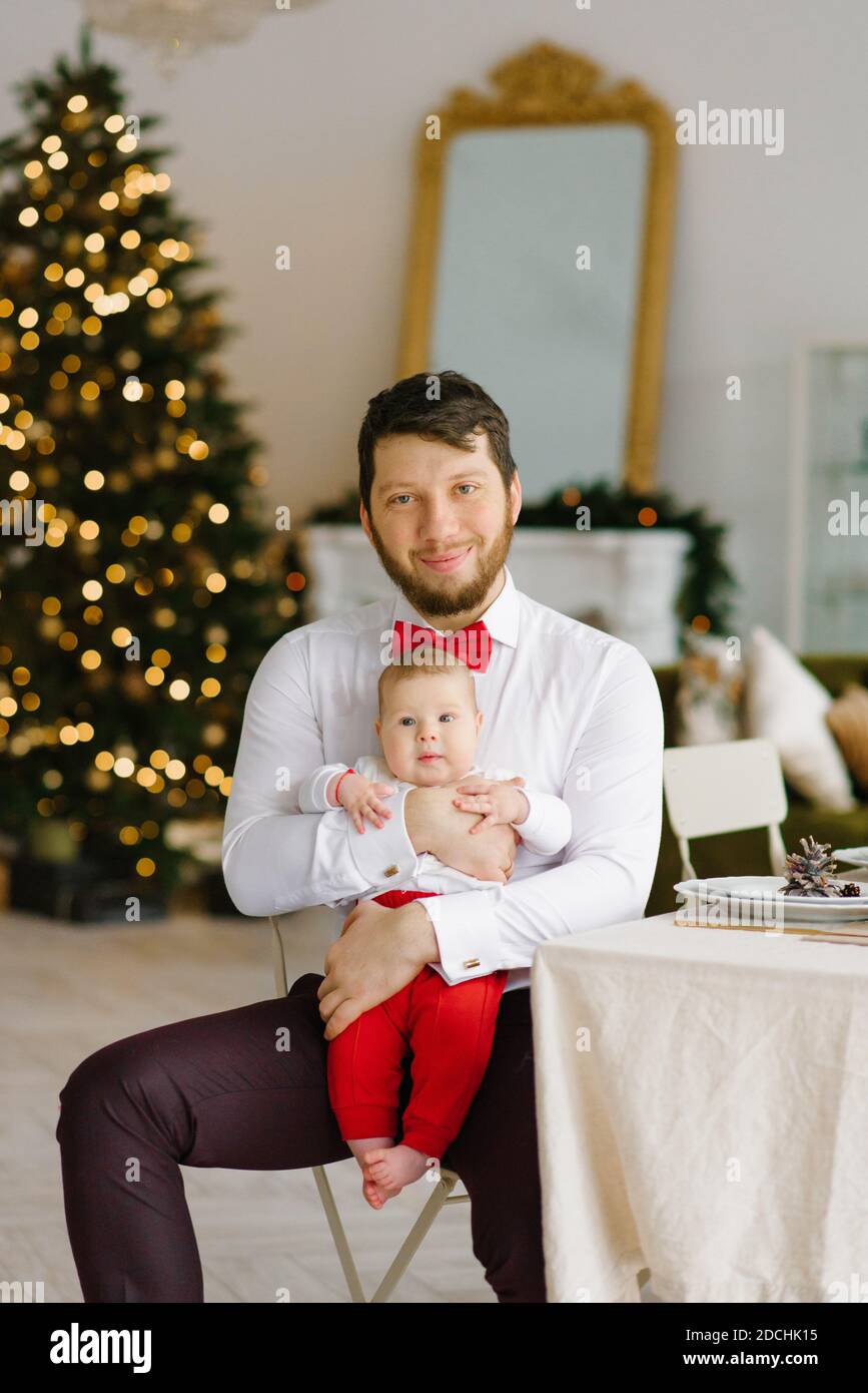 Un père élégant dans une chemise blanche avec un rouge noeud papillon tient  son fils dans ses bras contre le Arrière-plan des lumières de Noël dans le  salon Photo Stock - Alamy