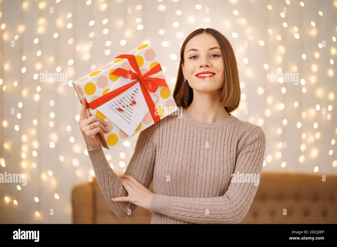 Portrait d'une jeune femme heureuse lèvres rouges tenant une boîte cadeau emballée. Gros plan la femme satisfaite a reçu la boîte actuelle. Arrière-plan des lumières de Noël festives. Banque D'Images