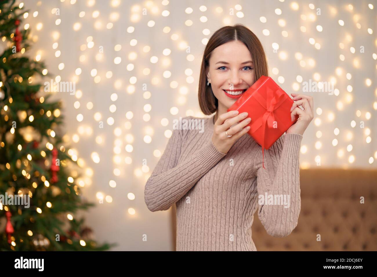 Portrait de jeune femme heureuse lèvres rouges regardant la boîte cadeau emballée. Gros plan la femme satisfaite a reçu la boîte actuelle. Arrière-plan des lumières de Noël festives Banque D'Images
