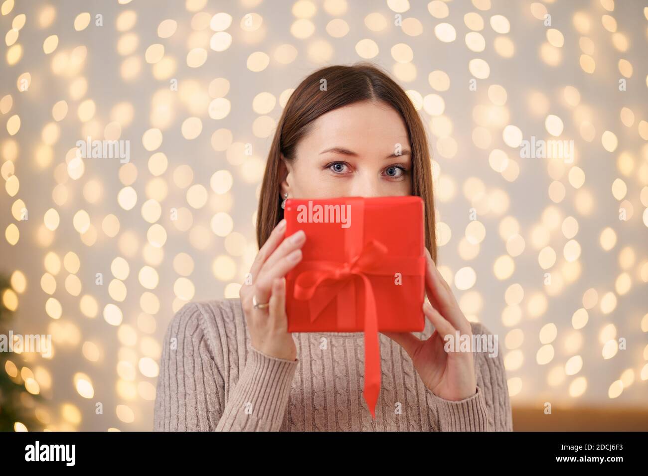 Portrait de la jeune femme heureuse lèvres rouges se cachant derrière la boîte cadeau emballée. Gros plan la femme satisfaite a reçu la boîte actuelle. Rétro-éclairage de Noël festif Banque D'Images