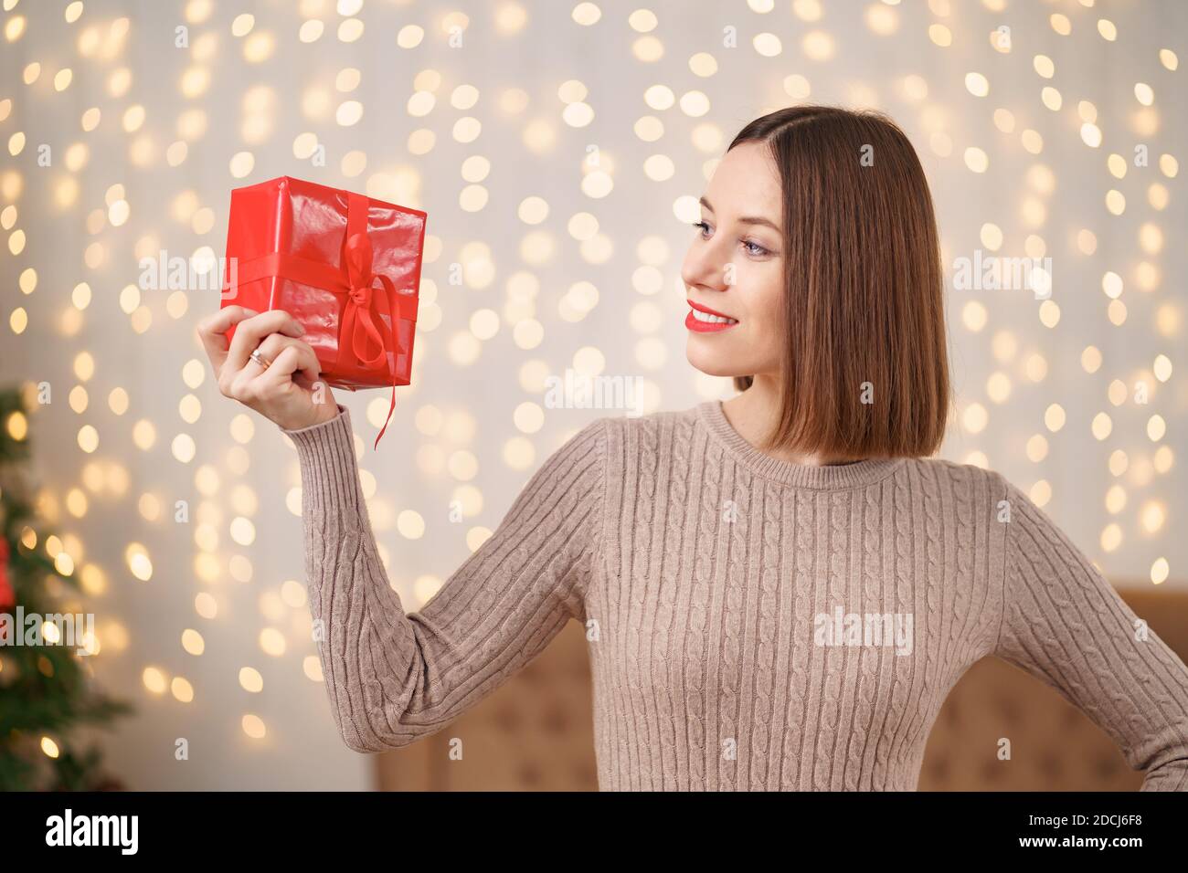 Portrait de jeune femme heureuse lèvres rouges regardant la boîte cadeau emballée. Gros plan la femme satisfaite a reçu la boîte actuelle. Arrière-plan des lumières de Noël festives Banque D'Images