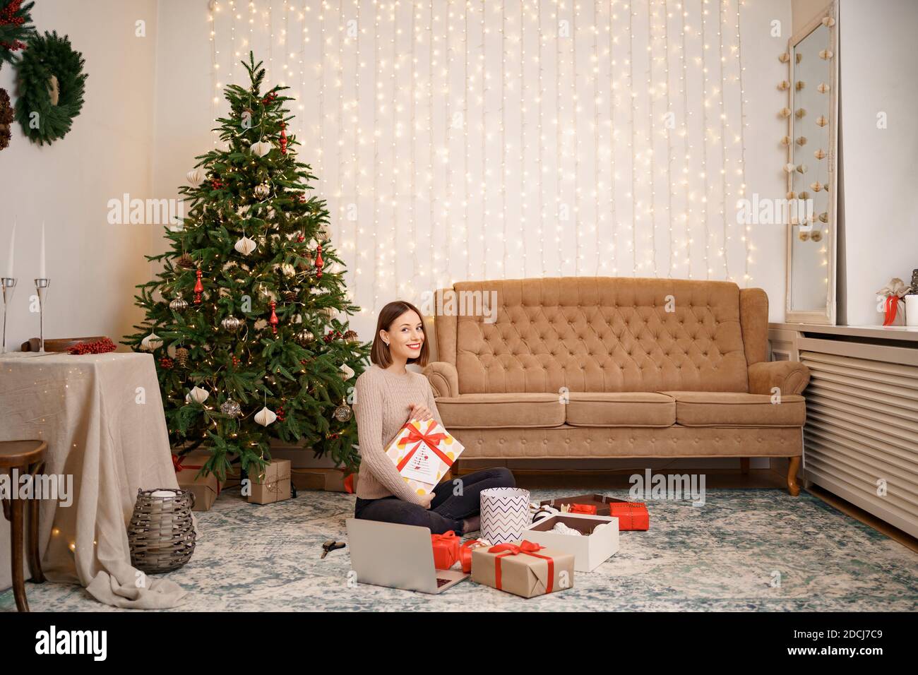 Portrait de jeune femme heureuse lèvres rouges tenir une boîte cadeau enveloppée assis entre beaucoup de cadeaux sur le tapis. Arrière-plan des lumières de Noël festives. Banque D'Images