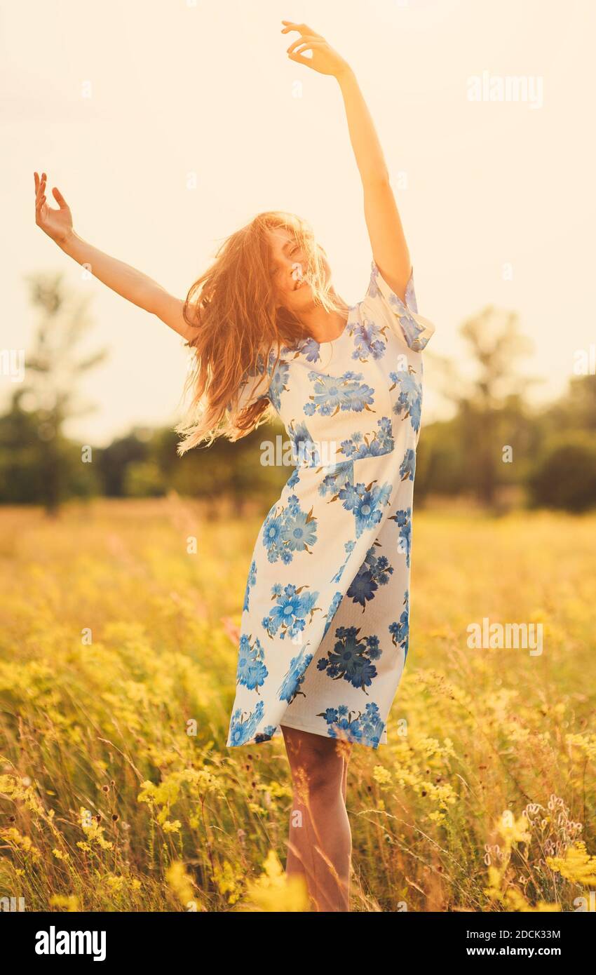 Belle tendre mignonne heureuse jeune fille jette ses mains belle blonde dans un champ de fleurs dans une robe multicolore sur fond de lumière de coucher de soleil Banque D'Images