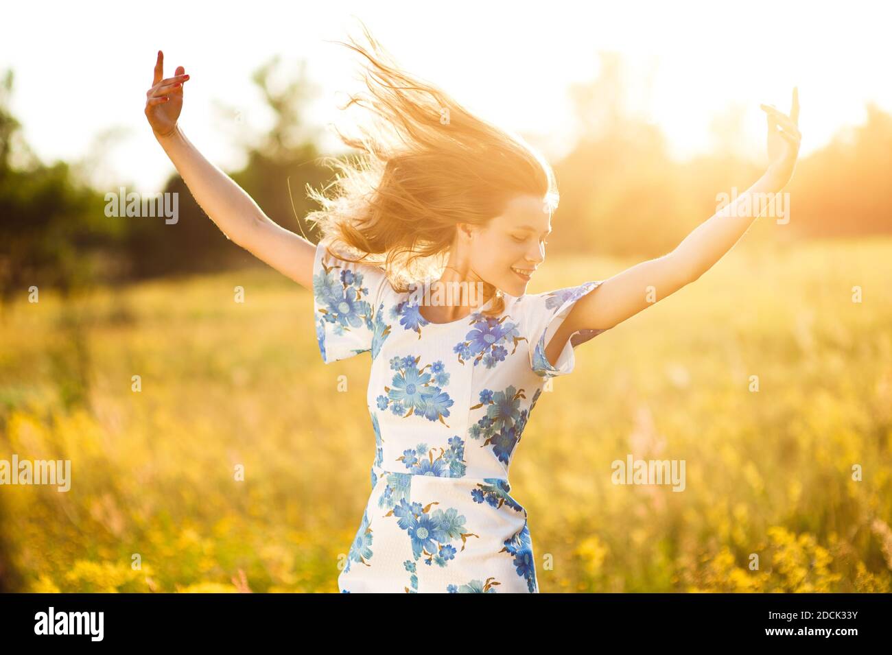 Belle tendre mignonne heureuse jeune fille jette ses mains belle blonde dans un champ de fleurs dans une robe multicolore sur fond de lumière de coucher de soleil Banque D'Images