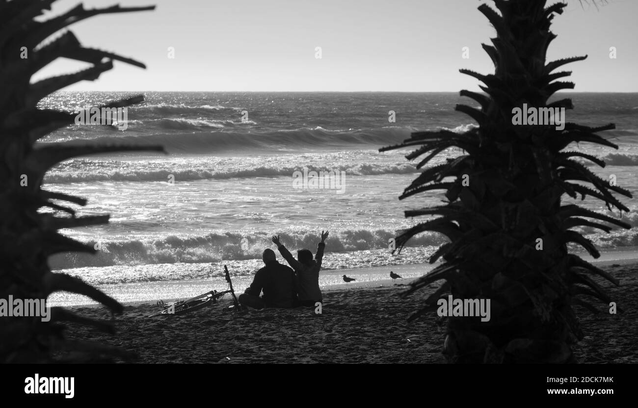 Femme excitée avec les mains dans l'air assis sur le sable à côté de l'ami sur la plage vide flanquée de palmiers à la Serena, au Chili. Photo en noir et blanc Banque D'Images