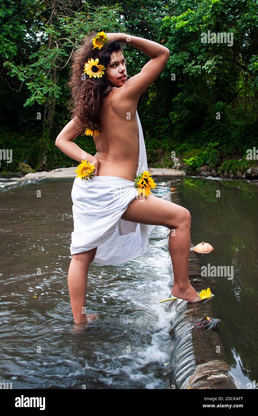Fille à la peau foncée et aux cheveux bouclés vêtue d'un tissu blanc et un torse nu entouré de tournesols et face peinte dans une rivière dans une forêt par jour Banque D'Images