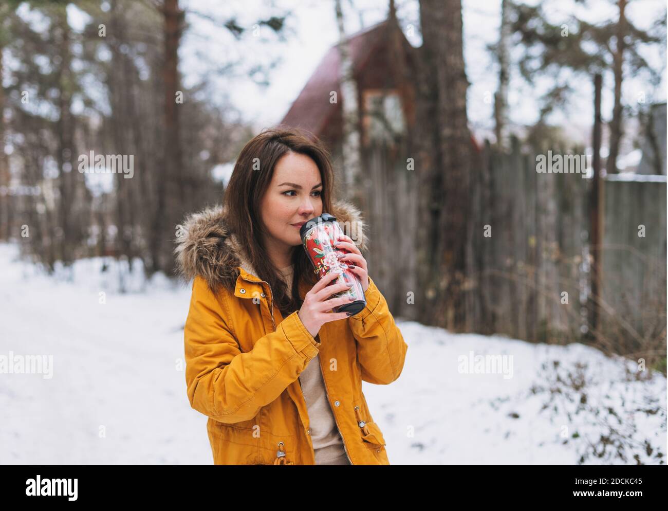 Jeune femme brune en blouson jaune avec mug thermo à petit village en forêt d'hiver Banque D'Images