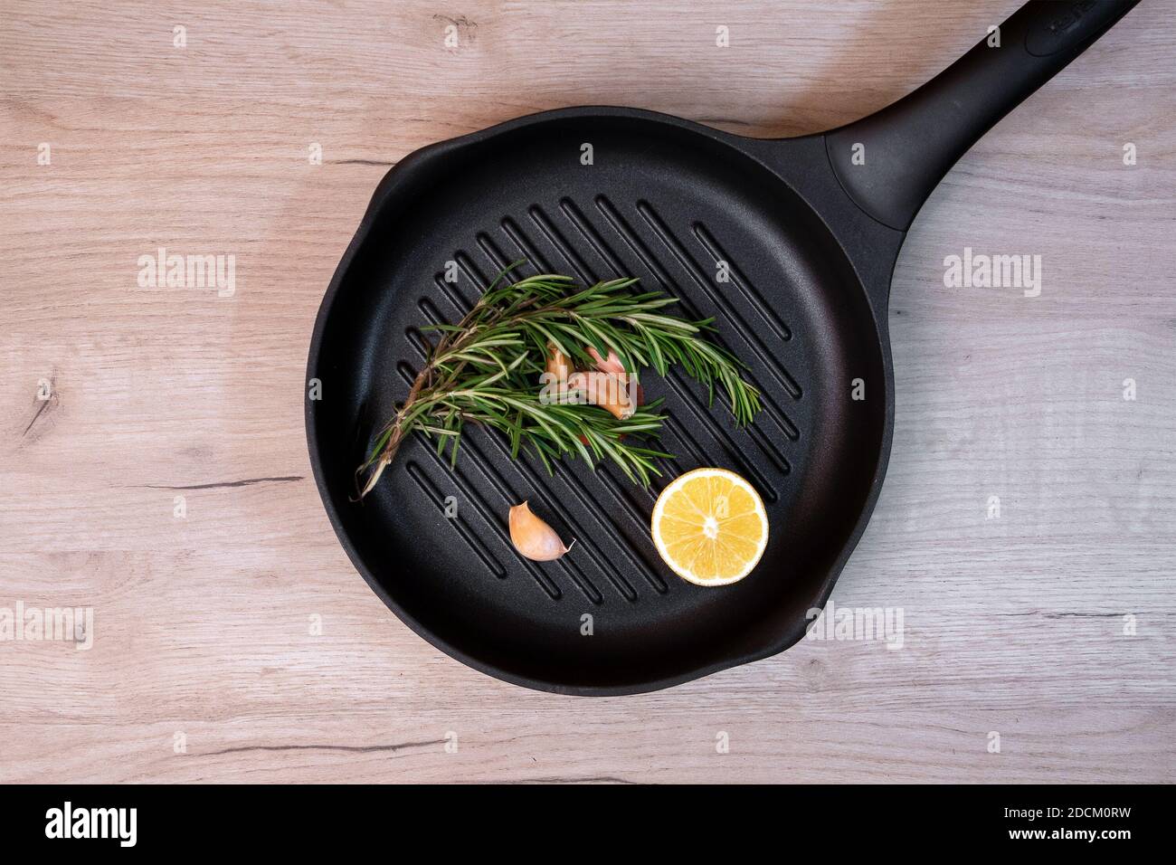Poêle à frire noire sur une planche de bois avec des branches de romarin, des gousses d'ail et une moitié de citron à l'intérieur Banque D'Images