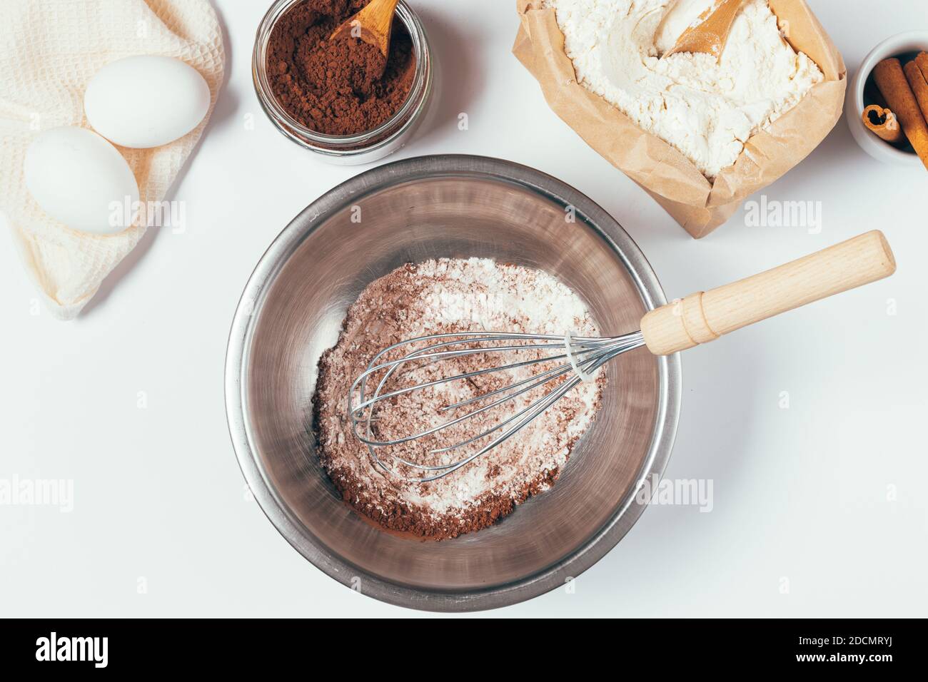 Bol avec mélange de farine et de poudre de cacao et fouet à côté des ingrédients pour cuire le gâteau au chocolat à la cannelle maison sur table blanche, vue du dessus. Banque D'Images