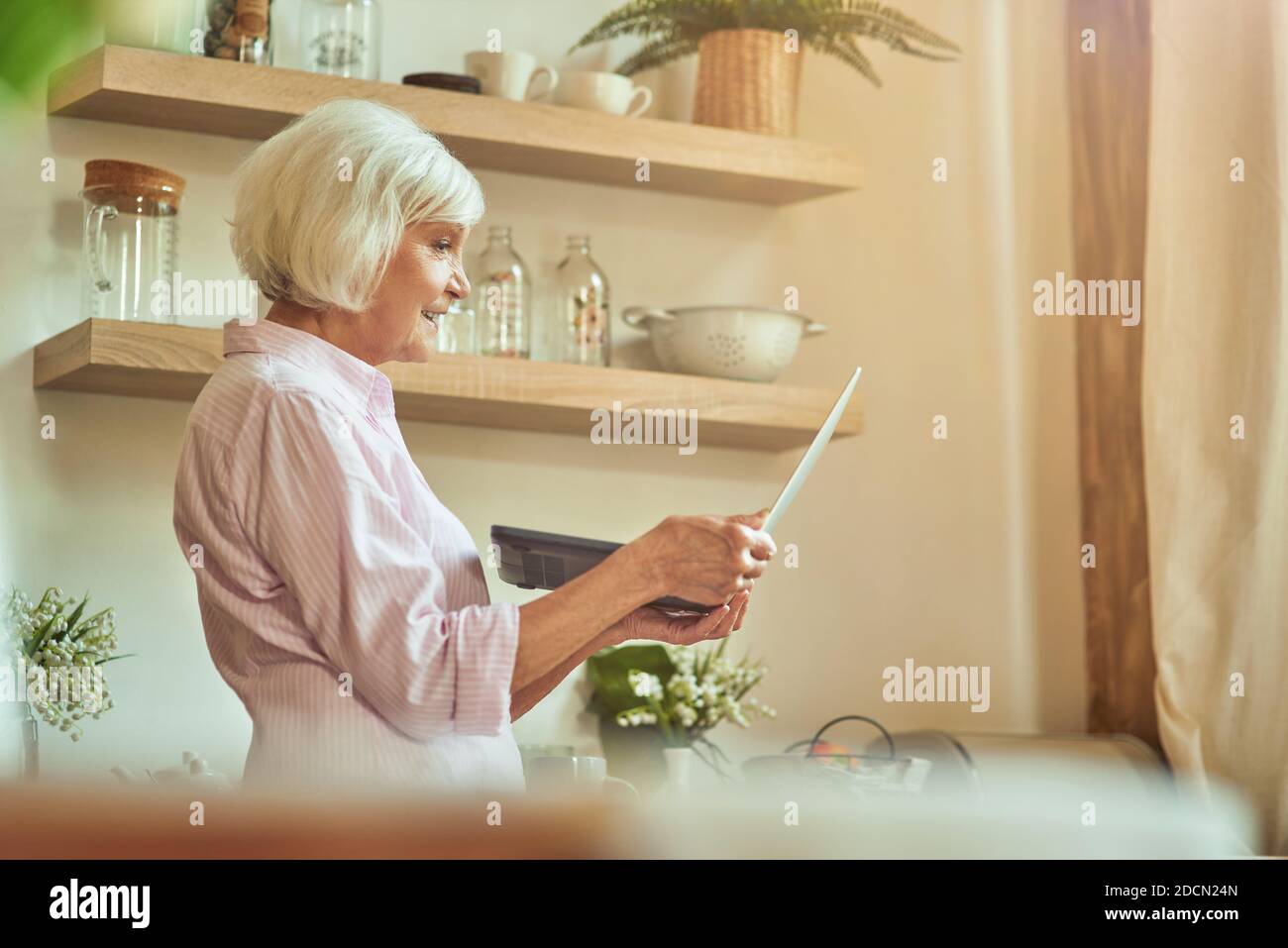 Vue latérale d'une femme âgée debout et tenant un ordinateur portable tout en regardant l'écran de la cuisine. Concept de style de vie domestique Banque D'Images