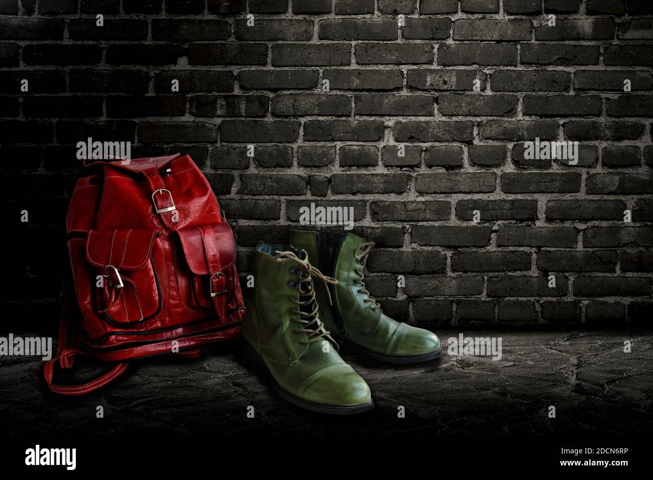 Bottes vert vintage et sac à dos rouge en cuir véritable sur fond de mur de briques sombres. Banque D'Images