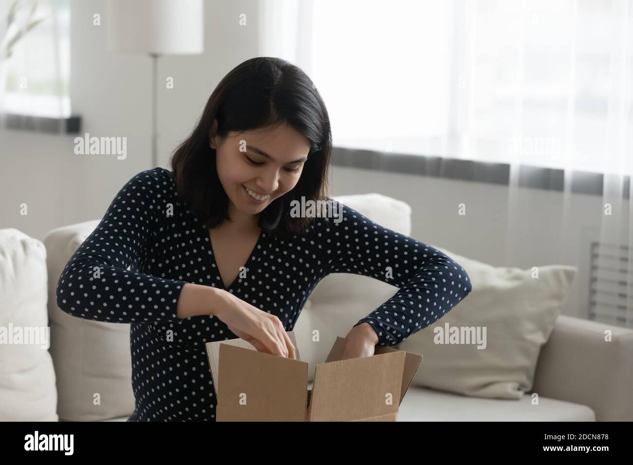 Femme asiatique souriante, en train de déballer un colis, assise sur un canapé à la maison Banque D'Images