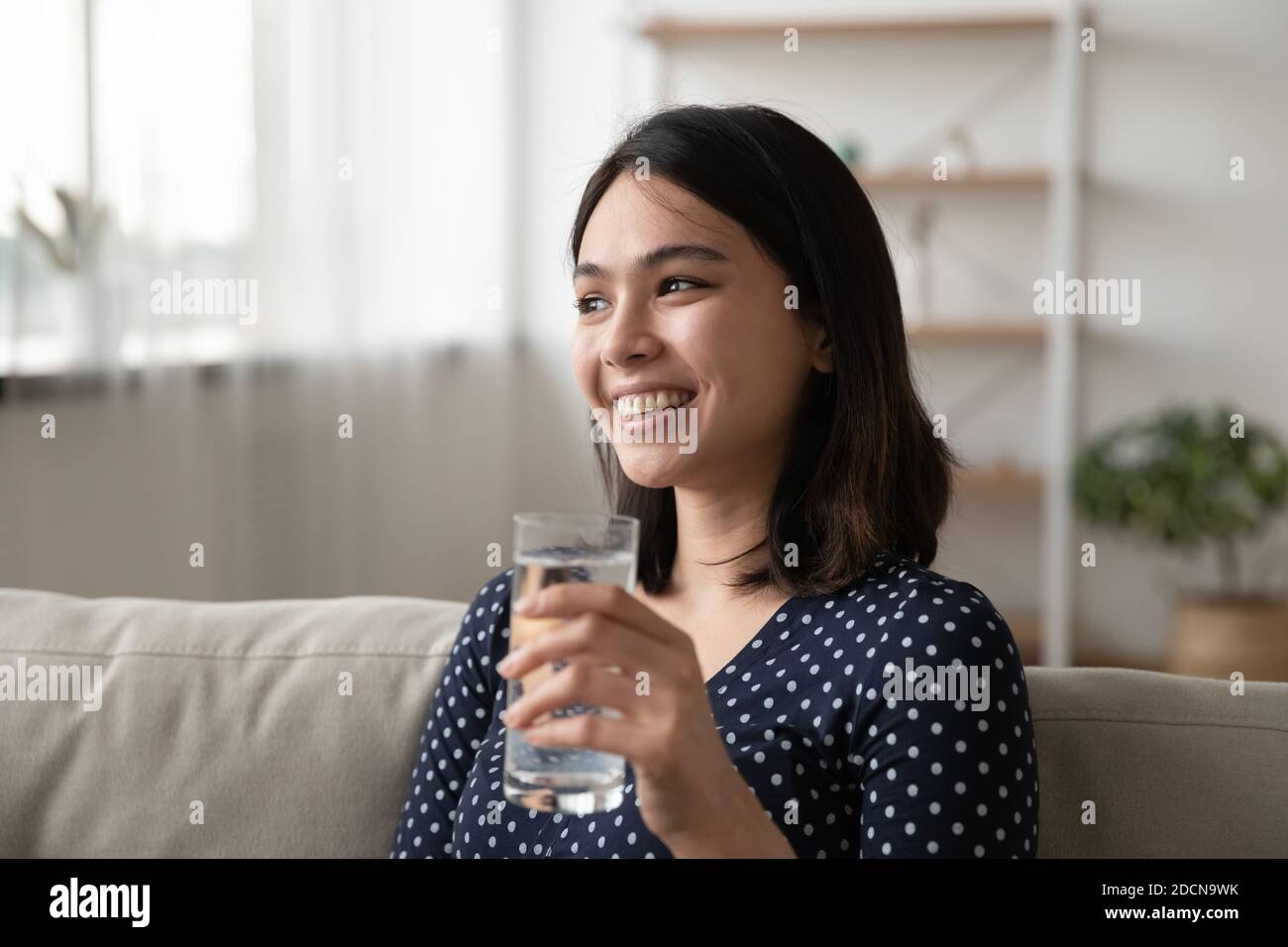 Gros plan femme asiatique souriante et rêveuse tenant un verre d'eau Banque D'Images
