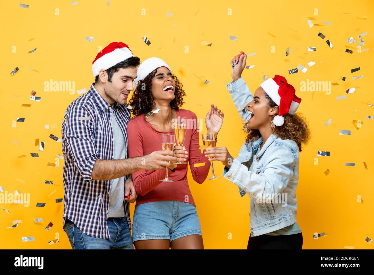 Groupe de trois heureux amis divers célébrant Noël boire du champagne sur fond jaune studio avec confetti Banque D'Images