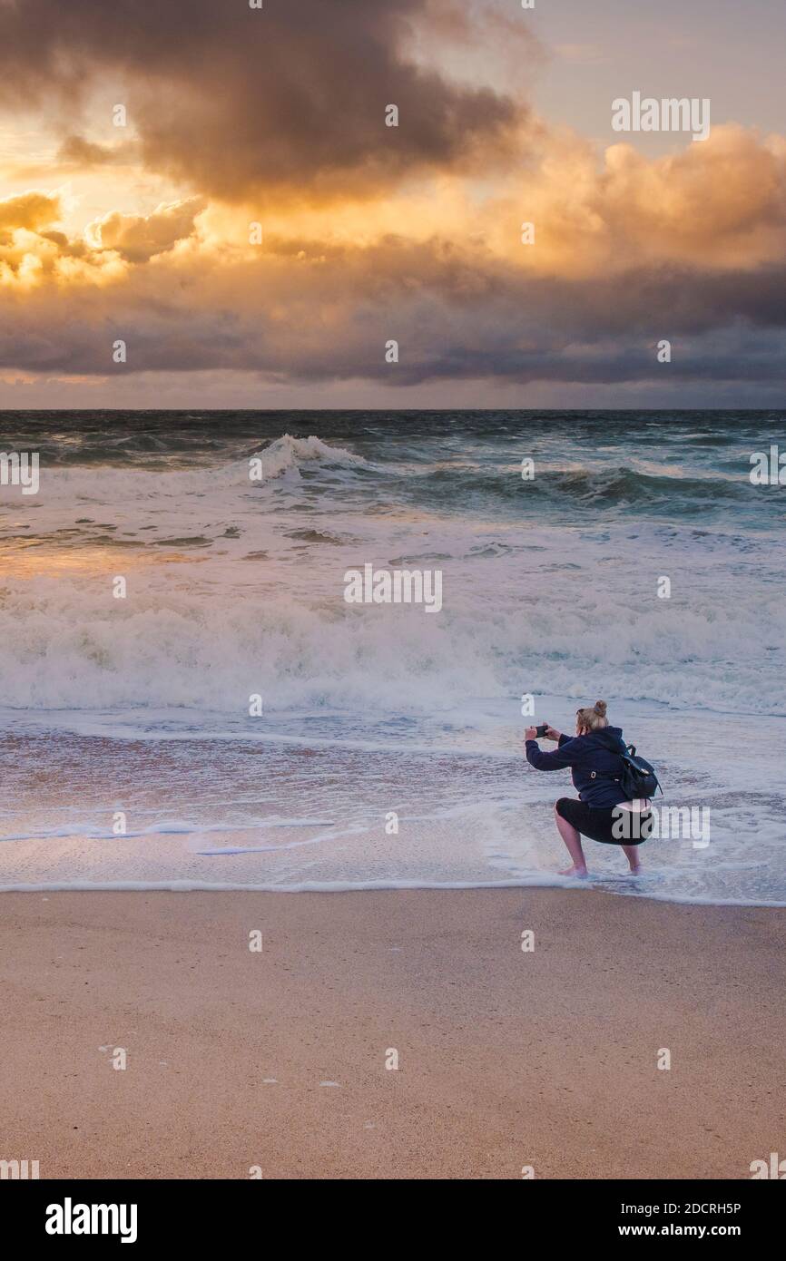 Un holidaymaker utilisant son téléphone portable pour photographier un coucher de soleil spectaculaire sur la plage de Fistral à Newquay en Cornouailles. Banque D'Images