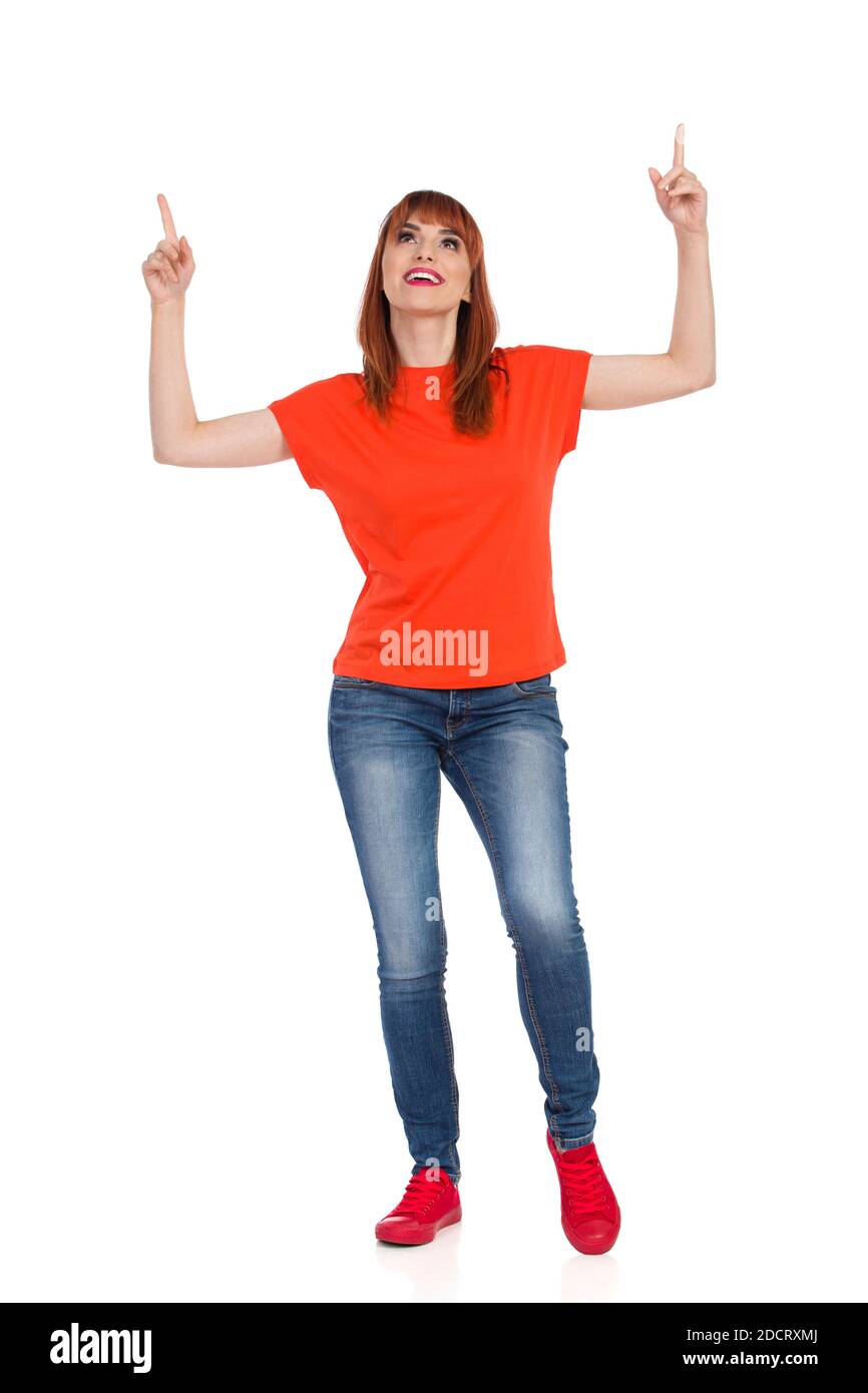 Décontracté jeune femme en chemise orange, jeans et baskets rouges est debout, regardant vers le haut, pointant et souriant. Vue avant. Prise de vue en studio pleine longueur isolée Banque D'Images