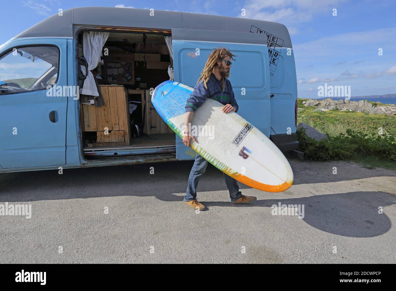 Un jeune surfeur mâle adulte en combinaison a mis hors planche à voile de la camionnette à Fistral Beach à Newquay , Cornwall, Royaume-Uni Banque D'Images