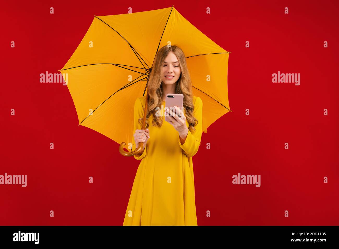 Belle jeune femme heureuse en robe jaune avec parapluie en utilisant téléphone mobile sur fond rouge Banque D'Images