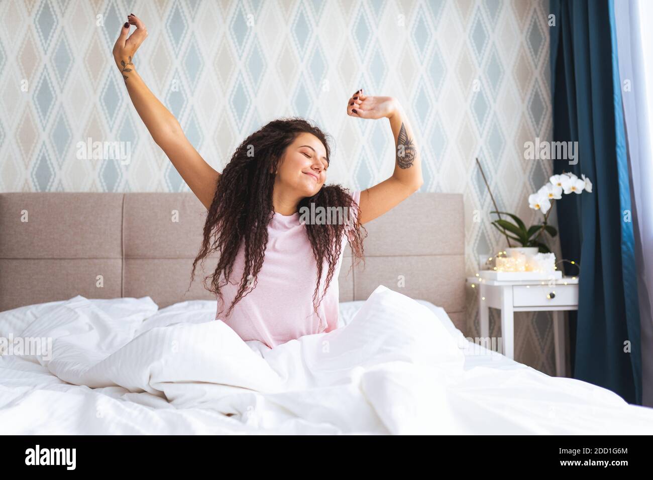 Vue en police de la jeune femme se réveillant tôt le matin assis sur le lit avec du linge blanc et de retcher ses bras et son corps avec le sourire. Banque D'Images