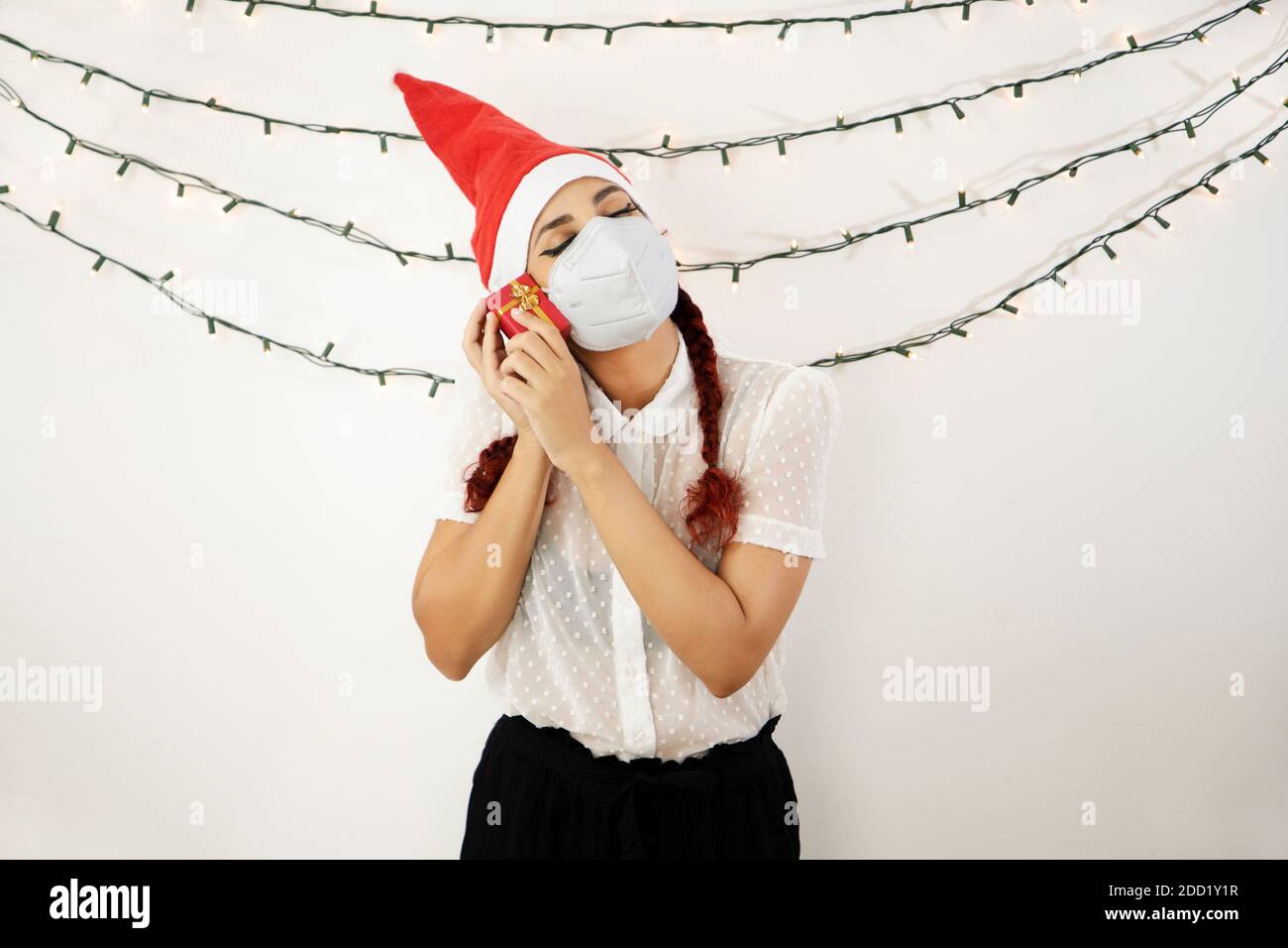 Fille avec un chapeau de Noël rouge et des cheveux rouges, avec un masque sur son visage collant une petite boîte cadeau rouge à son visage et fermant ses yeux. Belle fille Banque D'Images