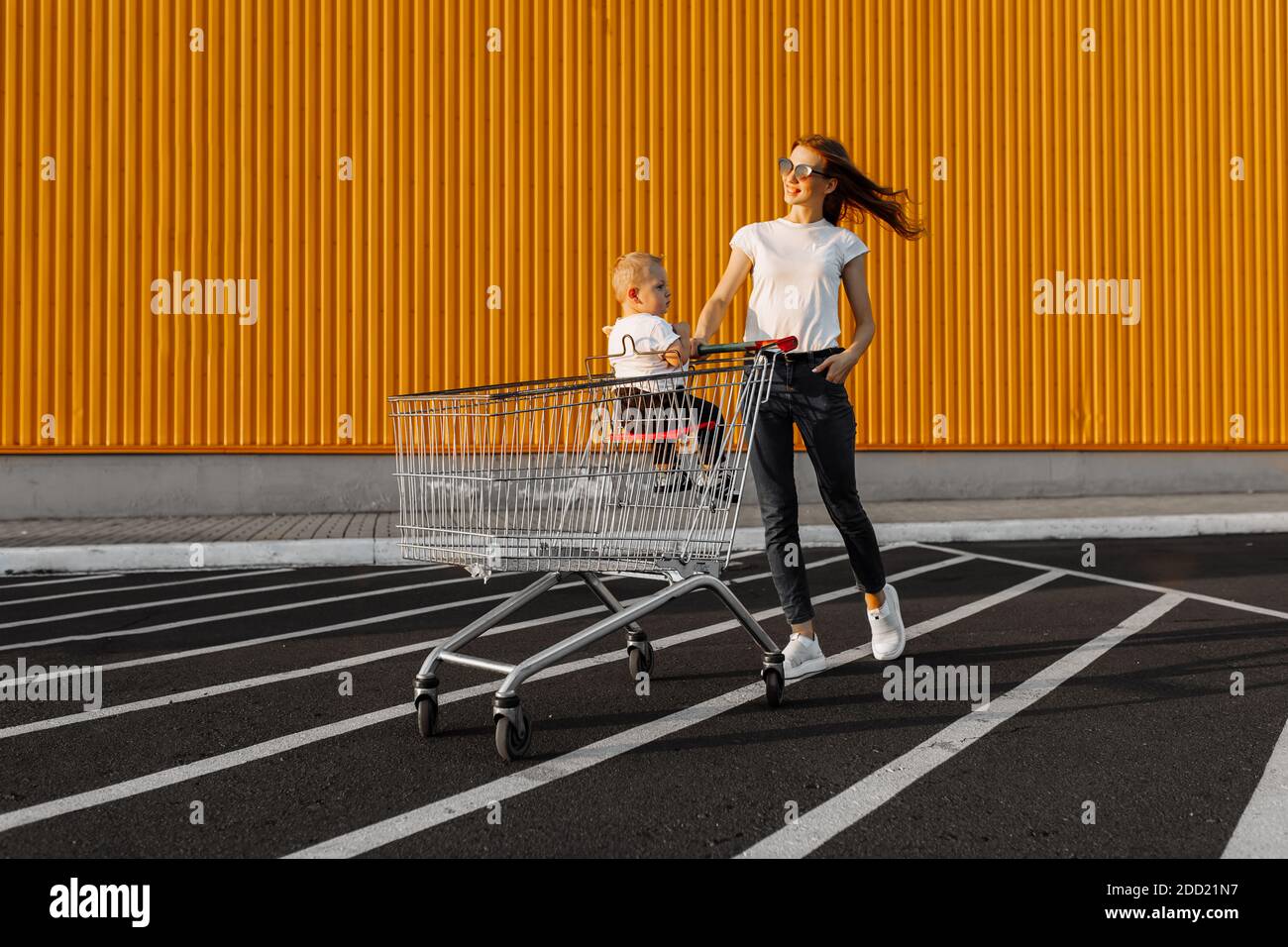 maman fait des achats avec un petit enfant assis dans un panier dans la rue sur le fond d'un mur jaune d'un centre commercial Banque D'Images