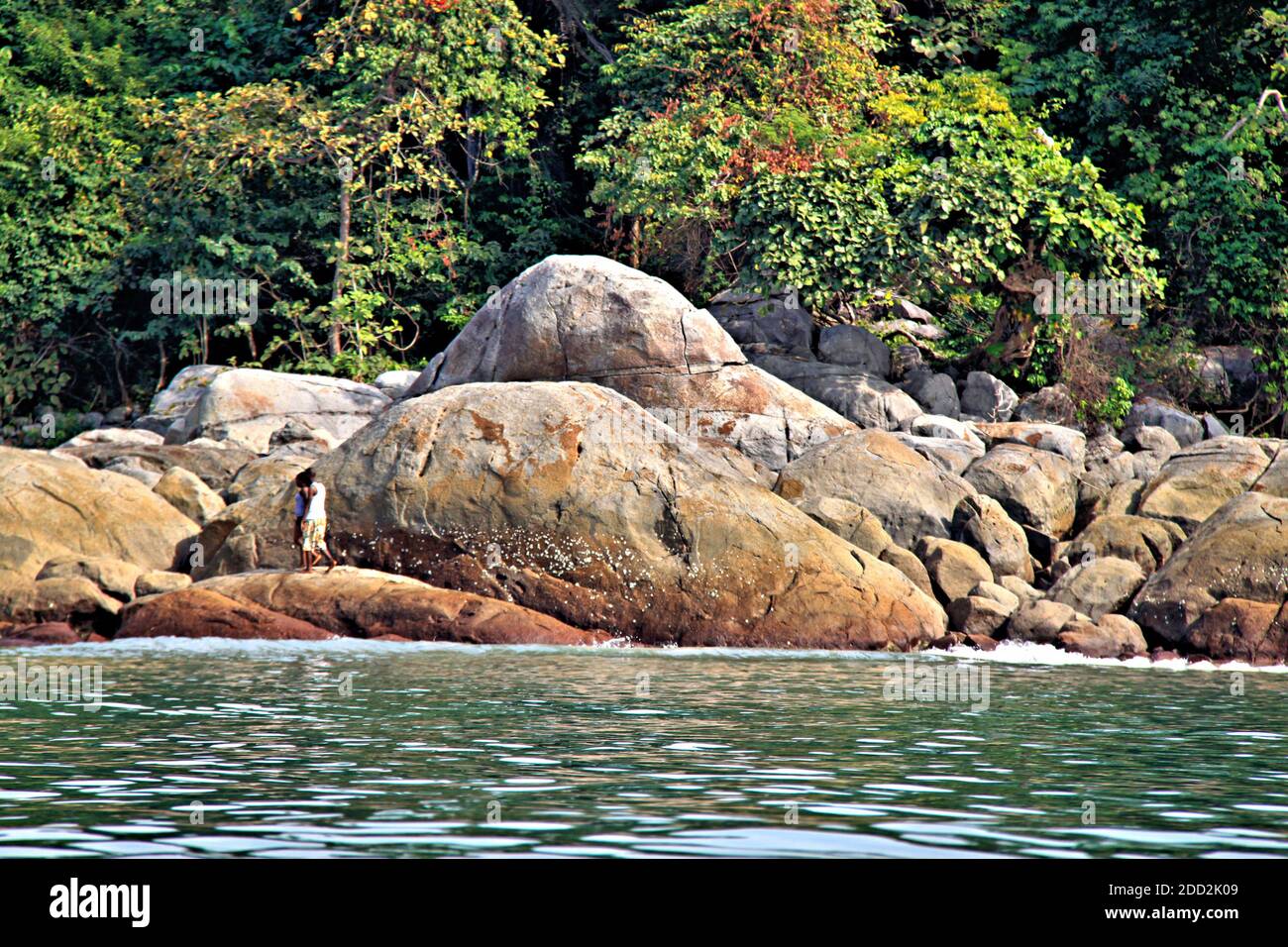 L'océan est au premier plan tandis que deux personnes marchent sur les grands rochers d'une île avec une forêt tropicale en arrière-plan. Banque D'Images