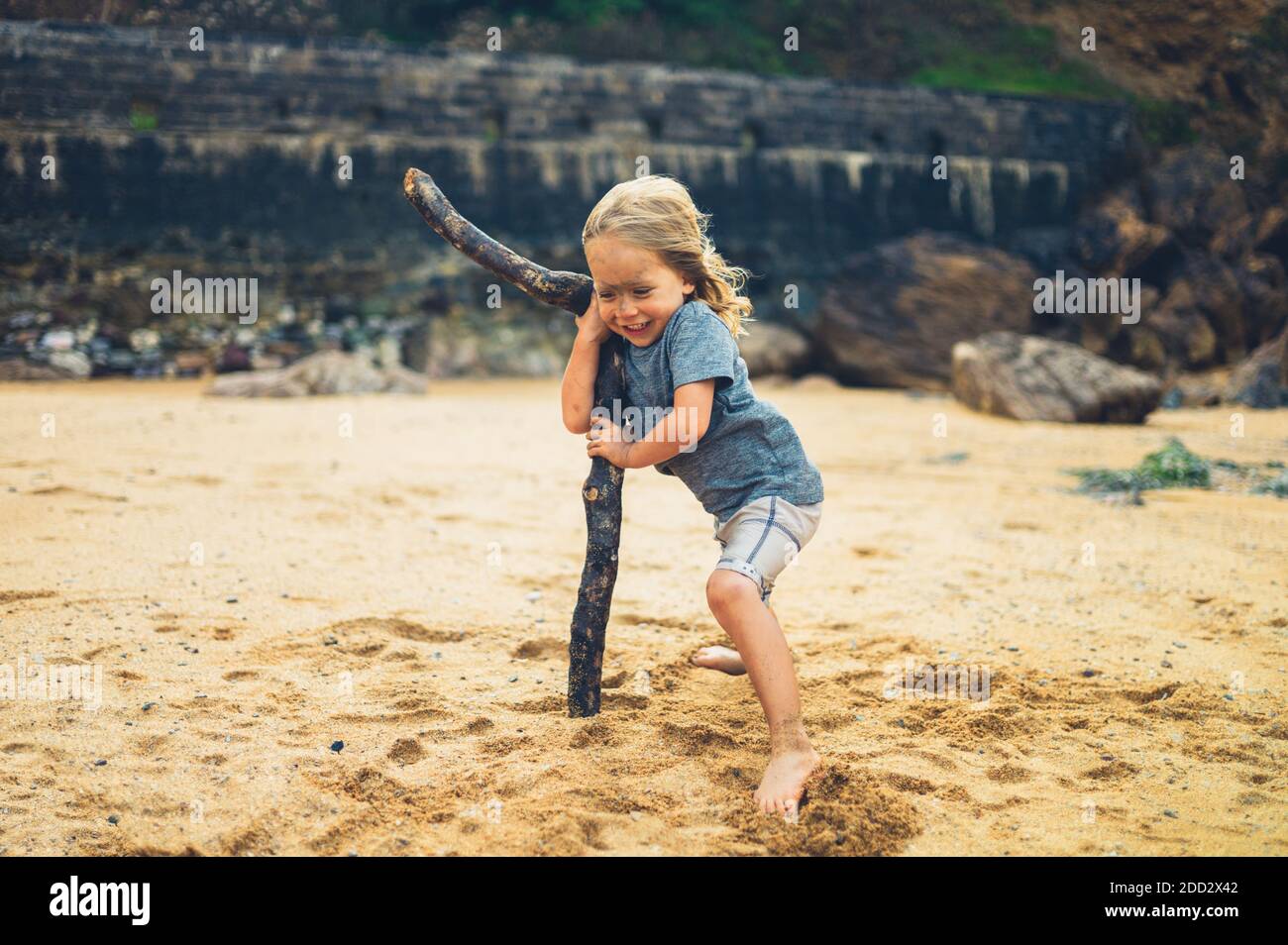 Un petit préchooler utilise un gros bâton pour soutenir lui-même contre le vent sur une plage Banque D'Images