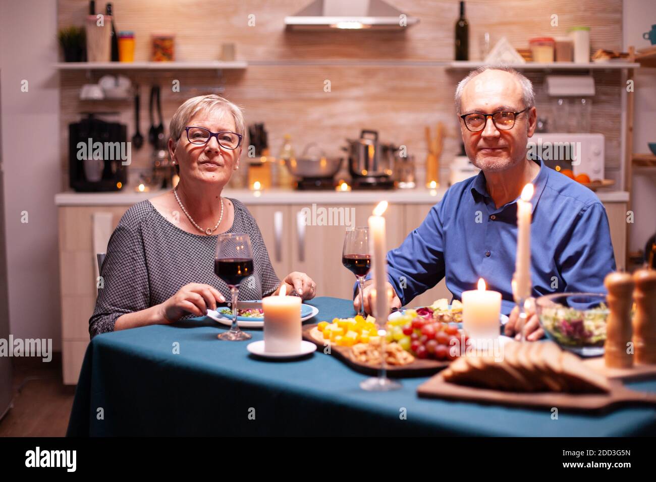 Joyeux couple dans la cuisine regardant l'appareil photo tenant des verres à vin regardant l'appareil photo pendant le dîner. Joyeux couple senior joyeux dînant ensemble dans la cuisine confortable, en appréciant le repas. Banque D'Images