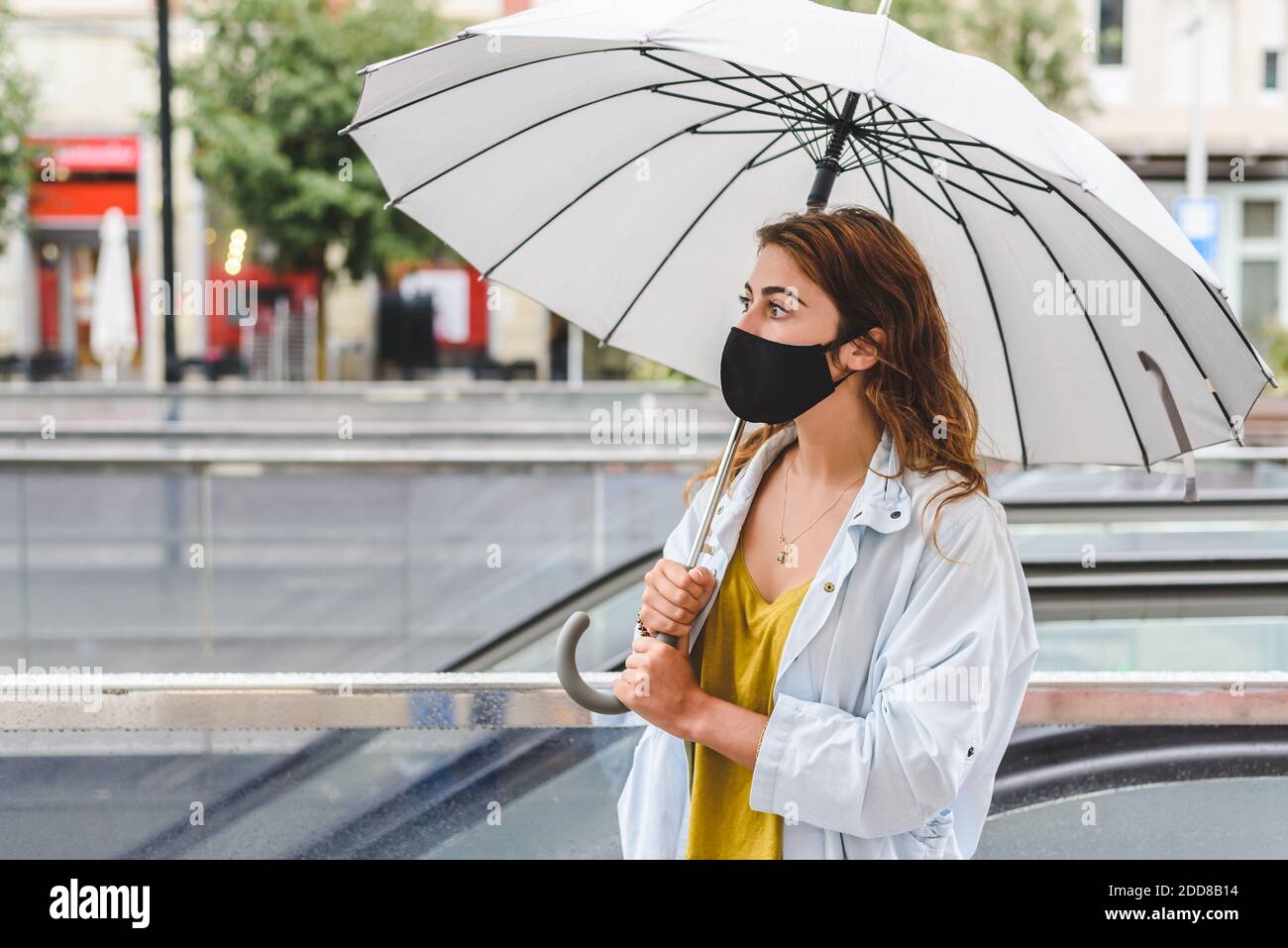 redhead jeune femme portant des vêtements décontractés et un visage noir masque en attente dans la rue près des escaliers souterrains et utilisation d'un parapluie sur une rai Banque D'Images