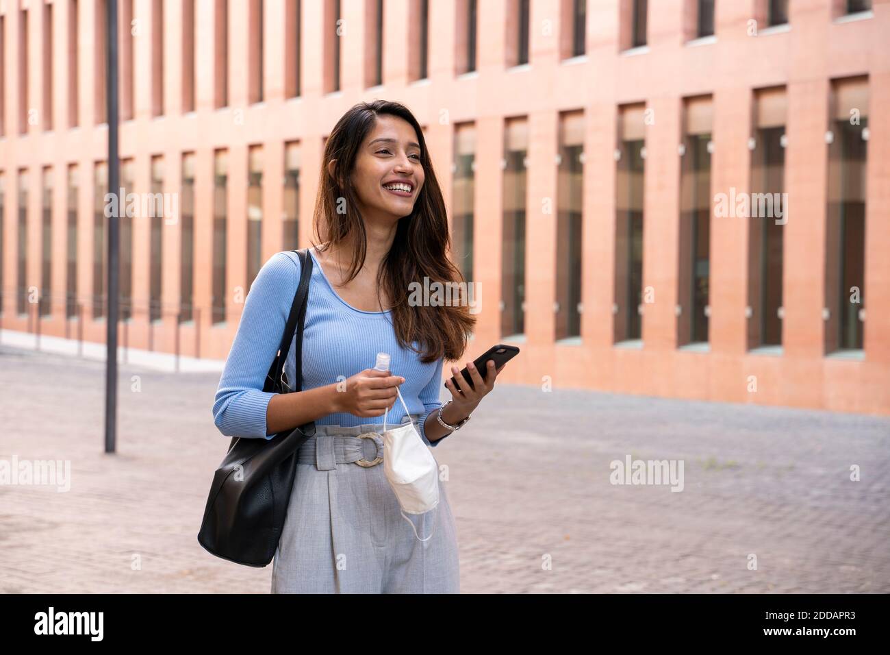 Jeune femme d'affaires souriante avec un smartphone tenant un masque de protection et de l'assainisseur pour les mains sur le chemin des pieds Banque D'Images