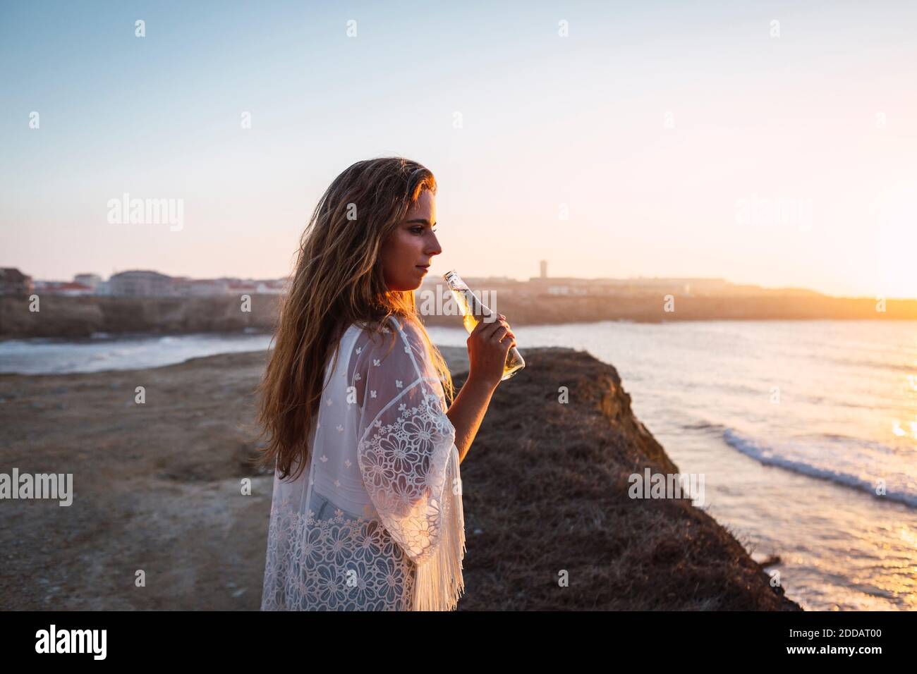 Jeune femme avec une petite bouteille de vin blanc qui regarde loin se tenir à la plage Banque D'Images