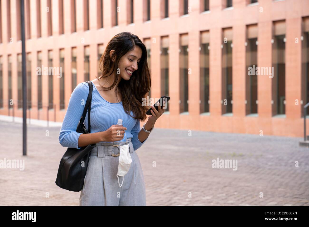 Jeune femme d'affaires souriante utilisant un smartphone tout en tenant le visage protecteur masque et désinfectant pour les mains sur le chemin du pied Banque D'Images