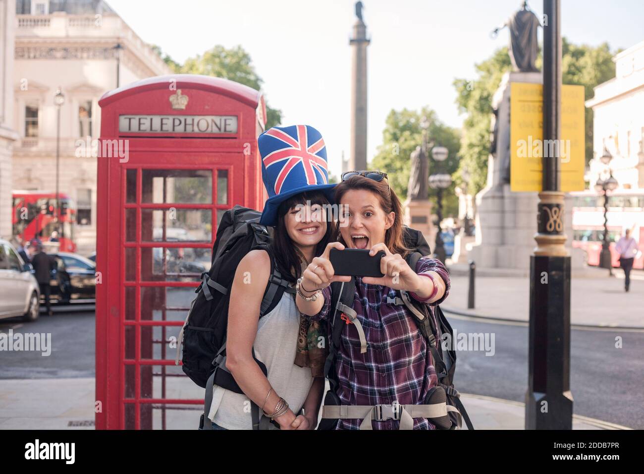 Une femme joyeuse prend un selfie avec un ami portant un chapeau drapeau britannique contre la boîte téléphonique rouge en ville Banque D'Images
