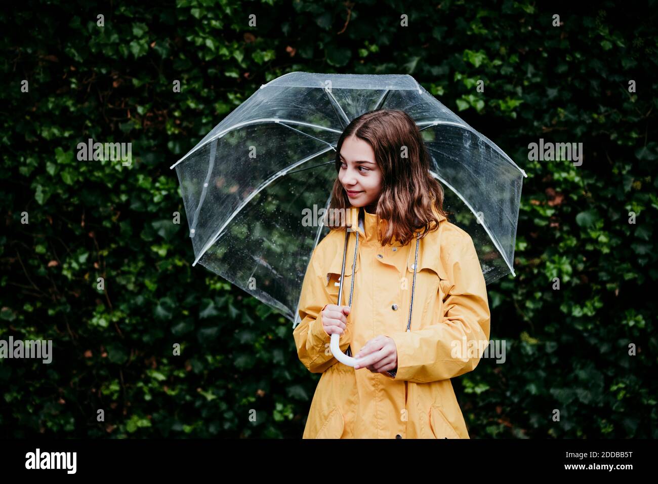 Fille portant un imperméable qui regarde loin tout en se tenant contre le mur de feuilles Banque D'Images