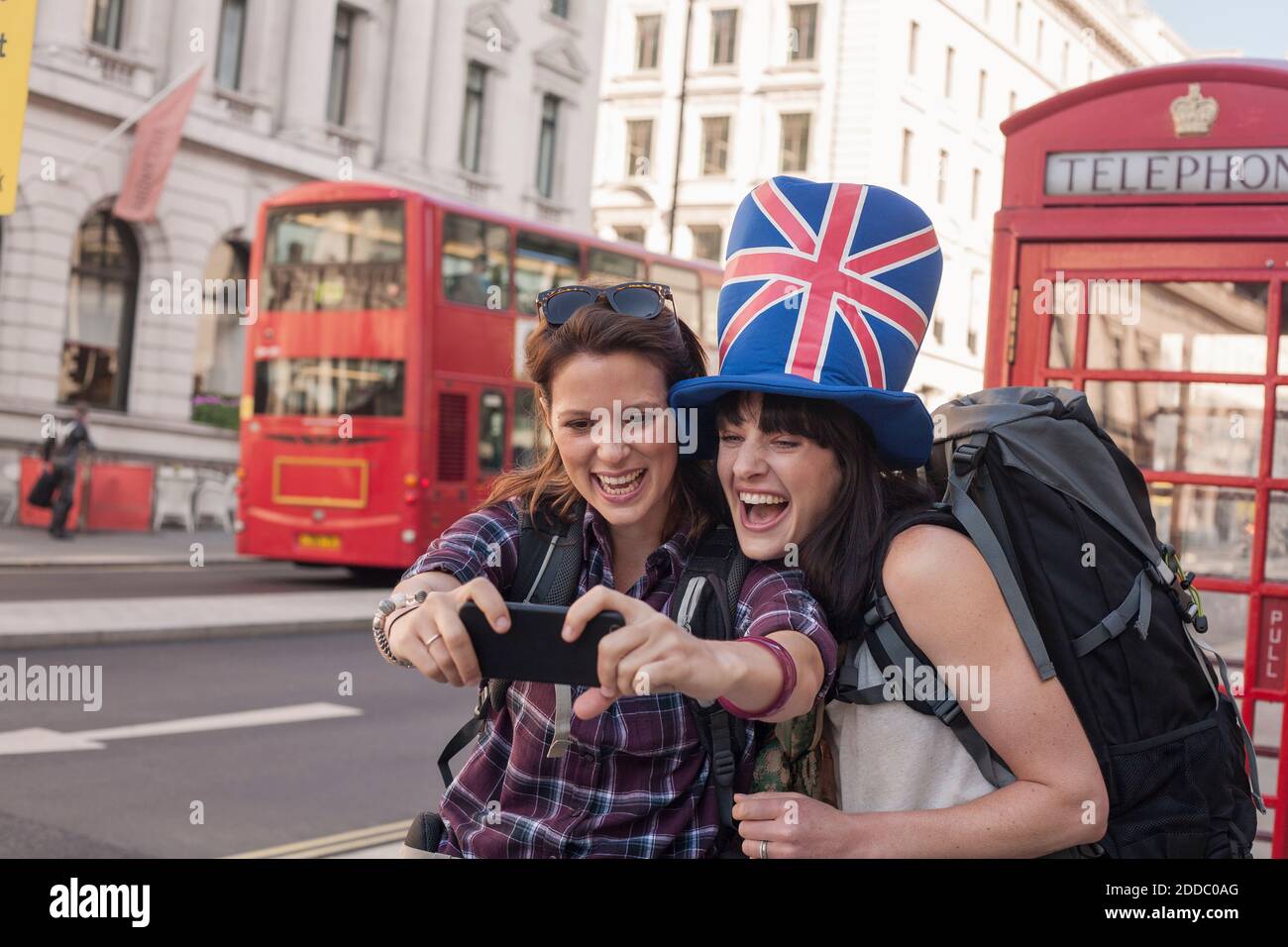 Bonne femme qui prend le selfie avec un ami portant un chapeau drapeau britannique contre la boîte téléphonique rouge en ville Banque D'Images