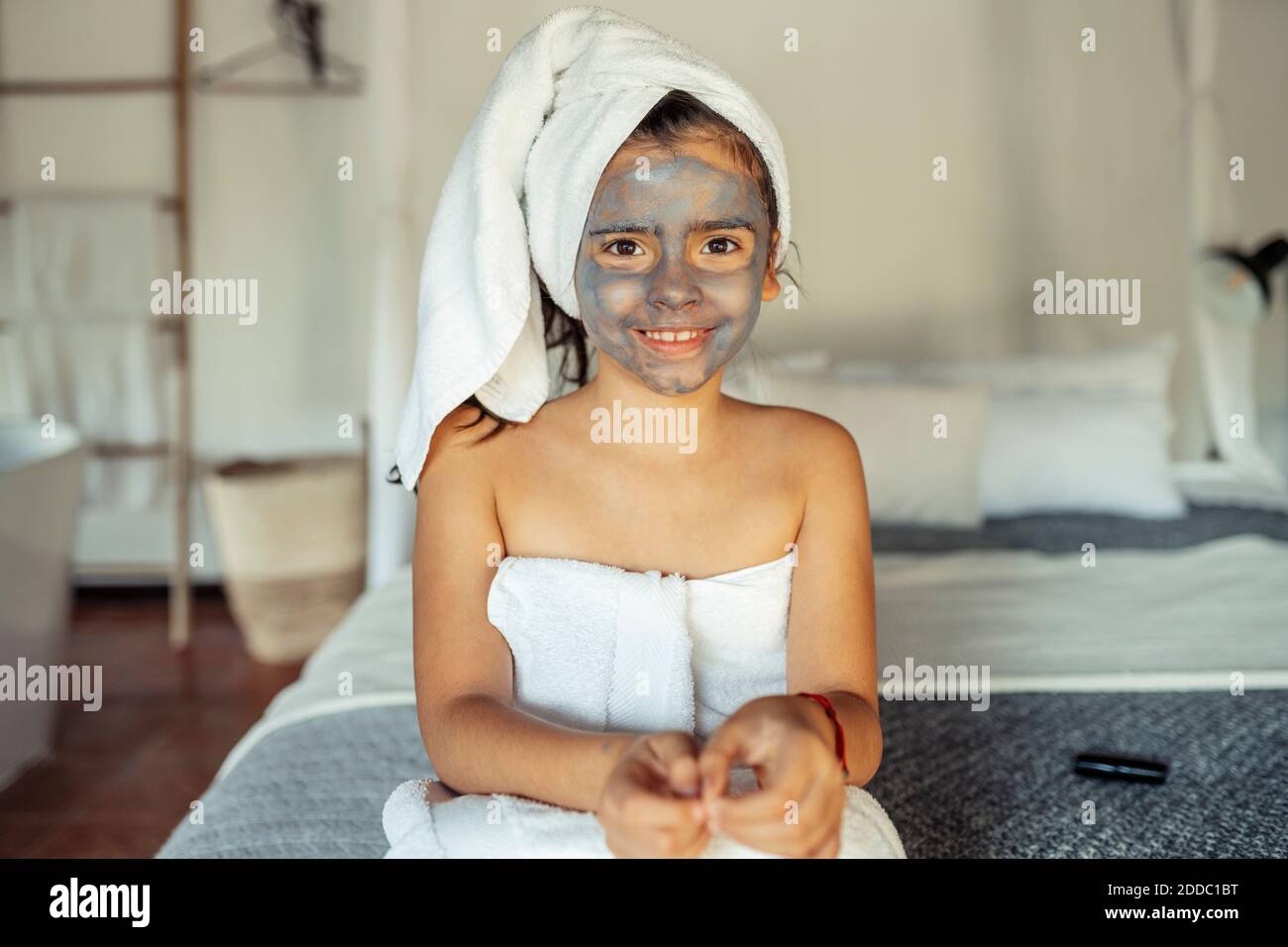 Fille souriante couverte d'une serviette avec masque facial sur le visage assis sur le lit à la maison Banque D'Images