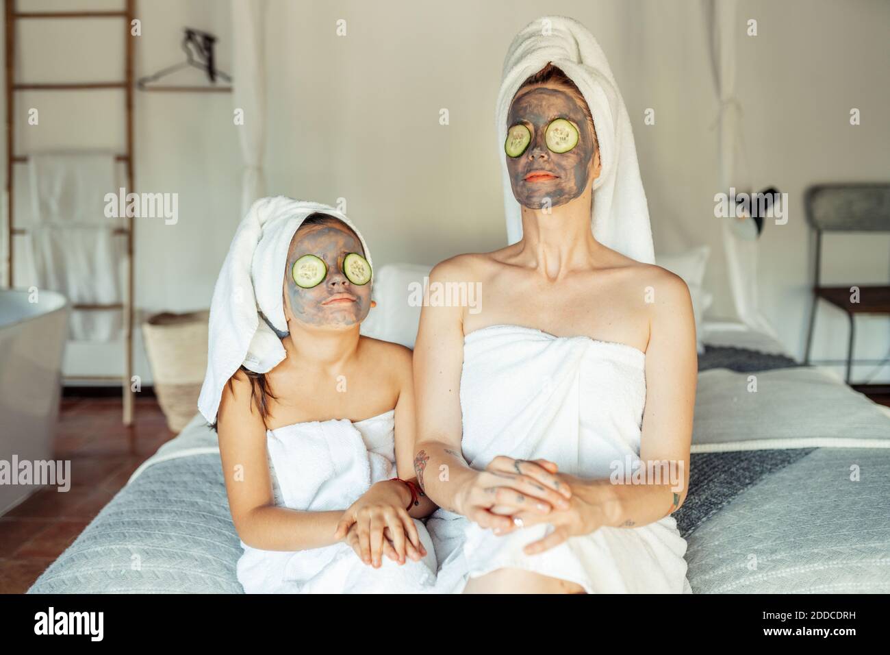Mère et fille dans une serviette avec masque facial et tranche de concombre sur les yeux assis à la maison Banque D'Images