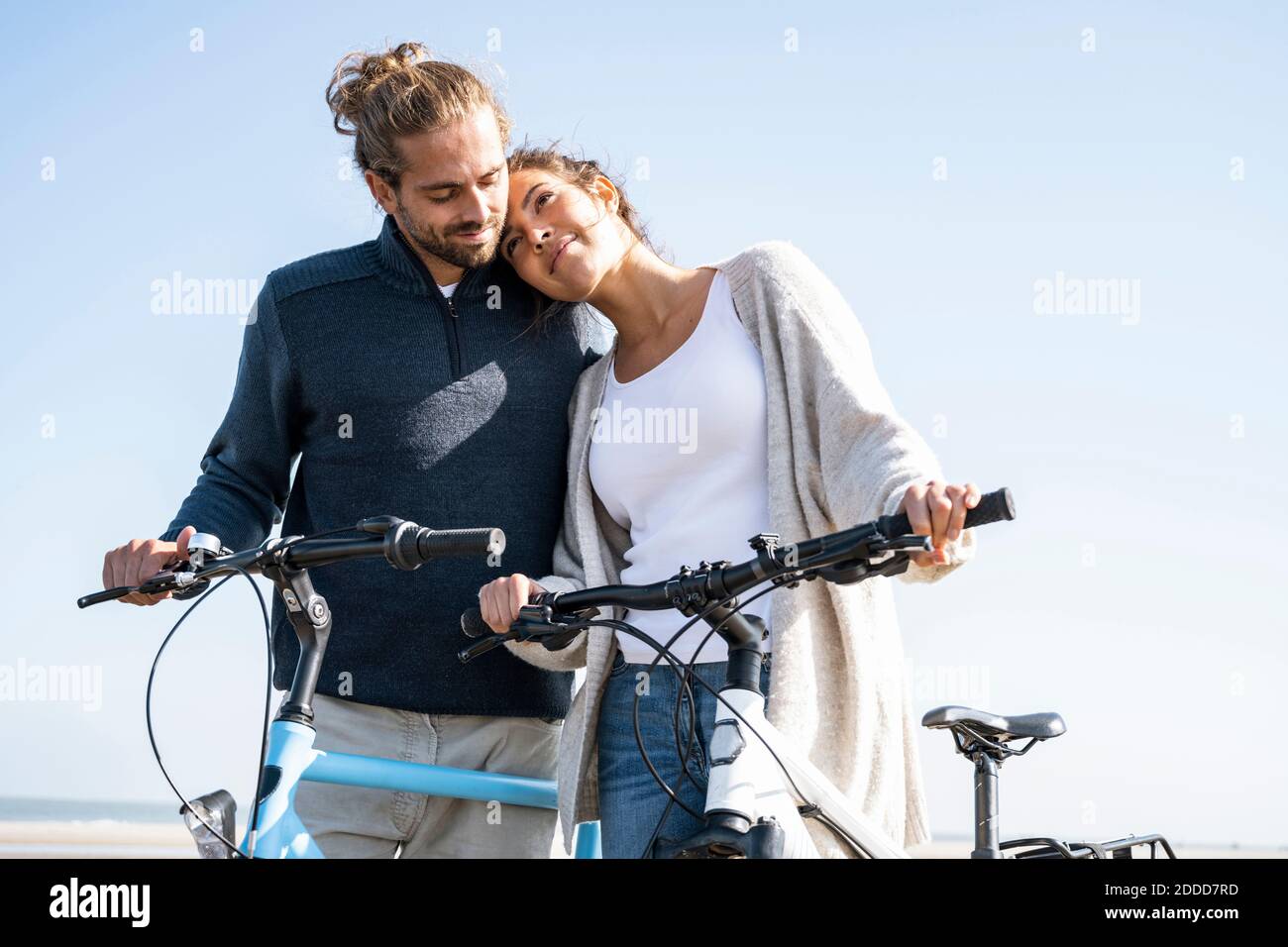 Belle femme avec la tête sur l'épaule de petit ami debout avec des vélos à la plage contre le ciel clair le jour ensoleillé Banque D'Images