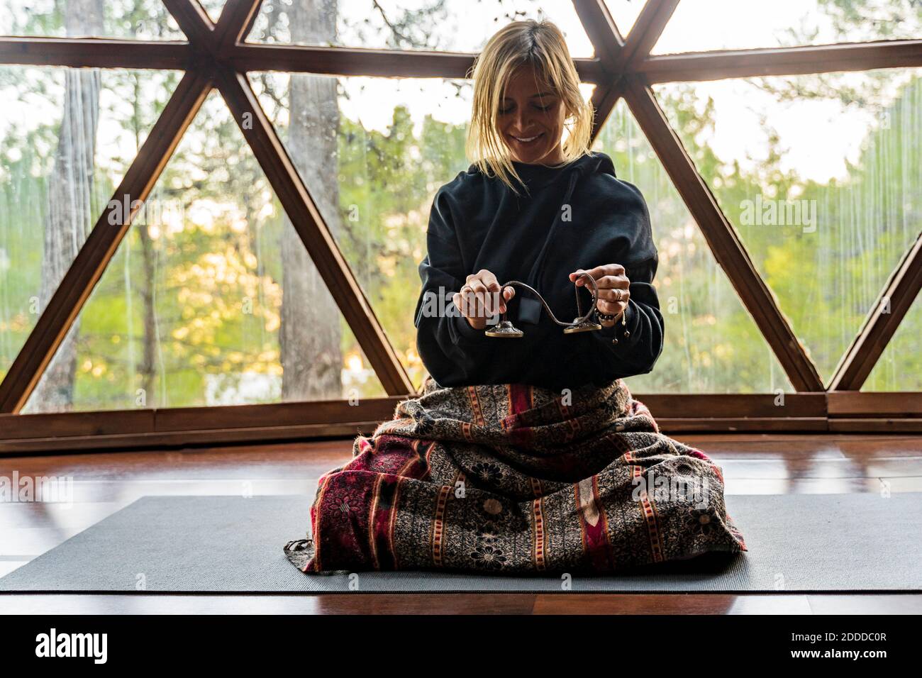 Femme blonde souriante instructeur de yoga tenant l'équipement tout en étant assise tapis contre fenêtre en classe d'exercice Banque D'Images