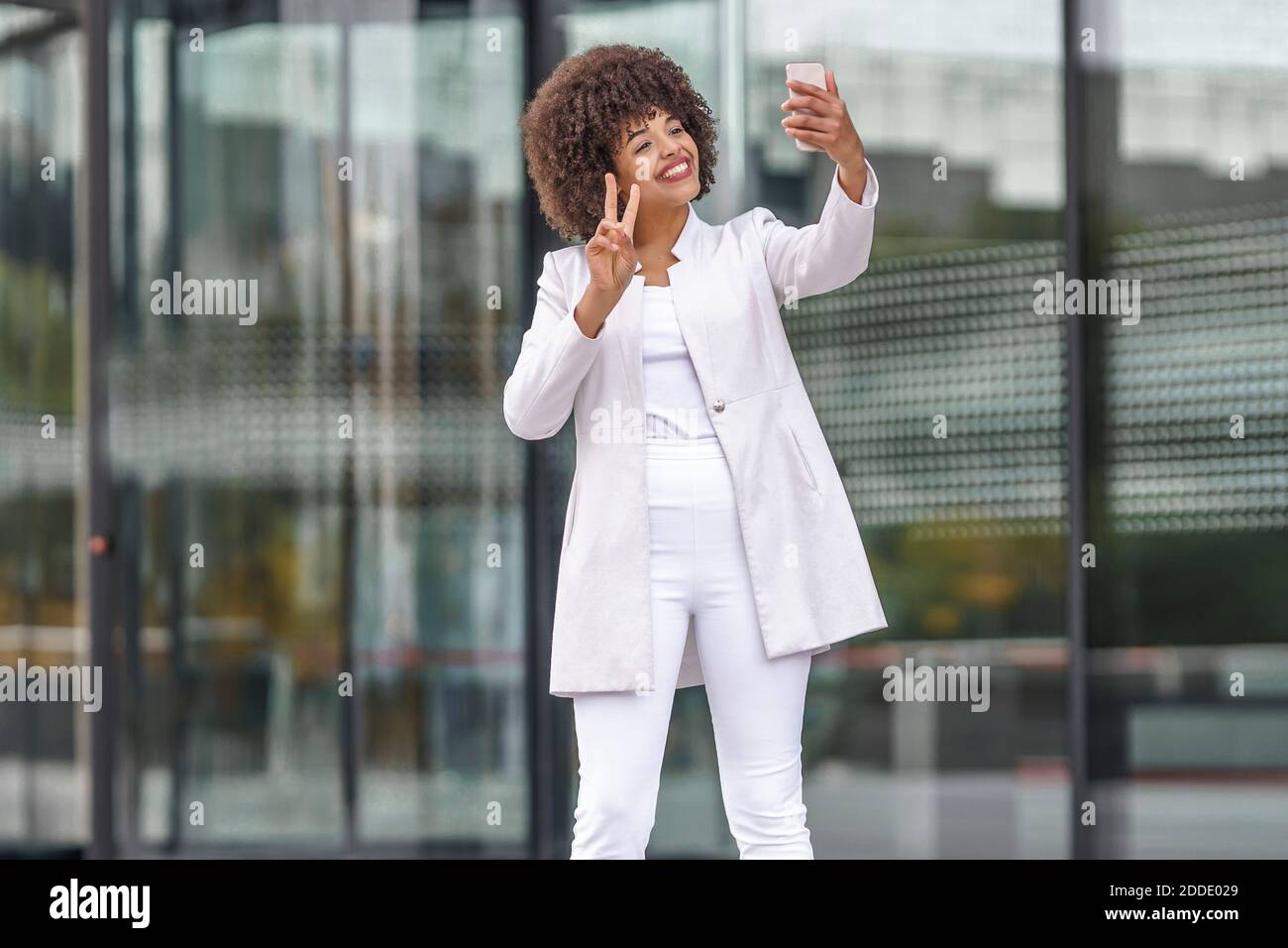 Femme d'affaires souriante montrant un geste de paix tout en prenant le selfie par mobile téléphone à l'extérieur Banque D'Images