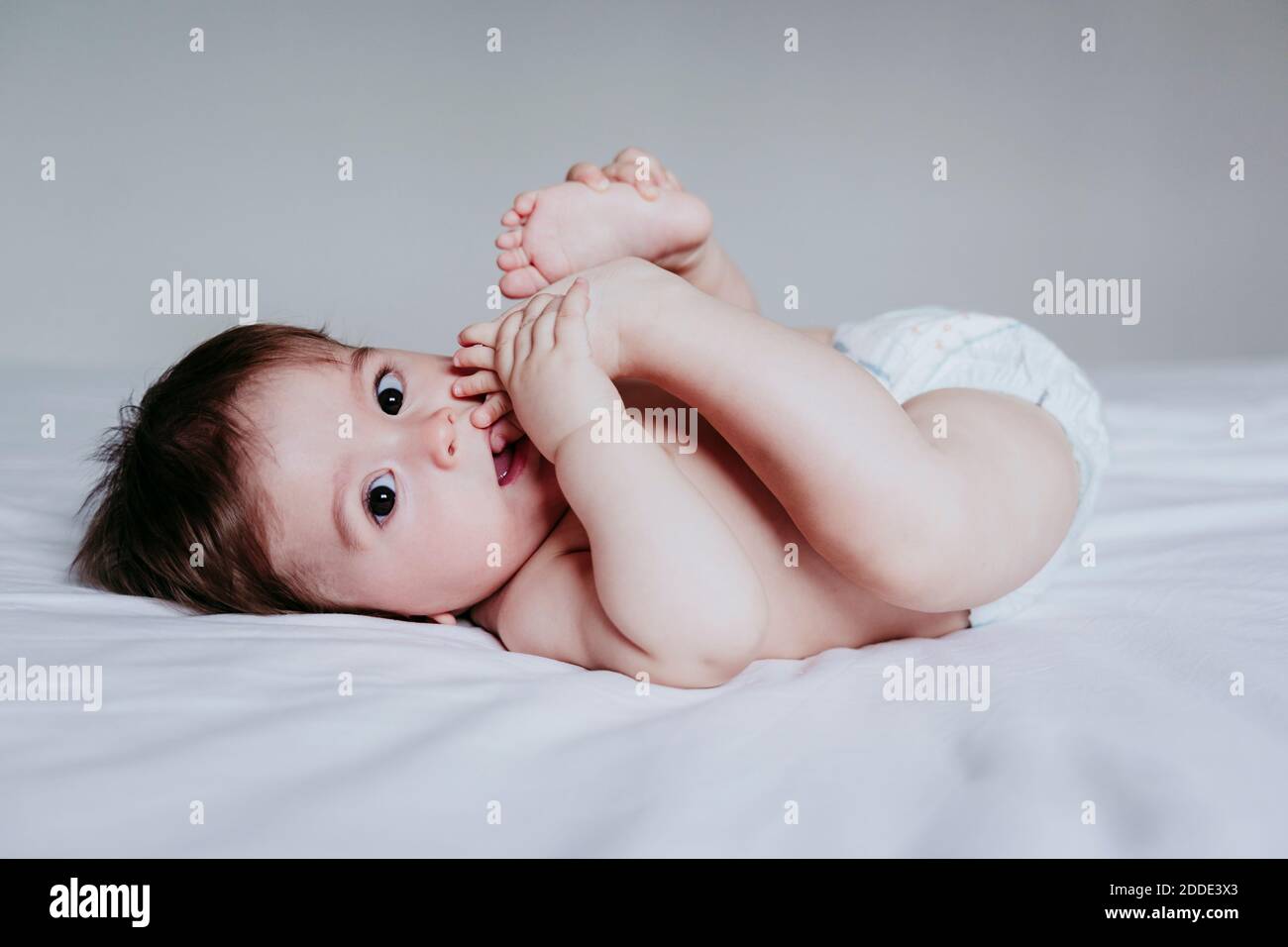 Bébé garçon mignon mettant le pied dans la bouche tout en étant allongé sur lit à la maison Banque D'Images