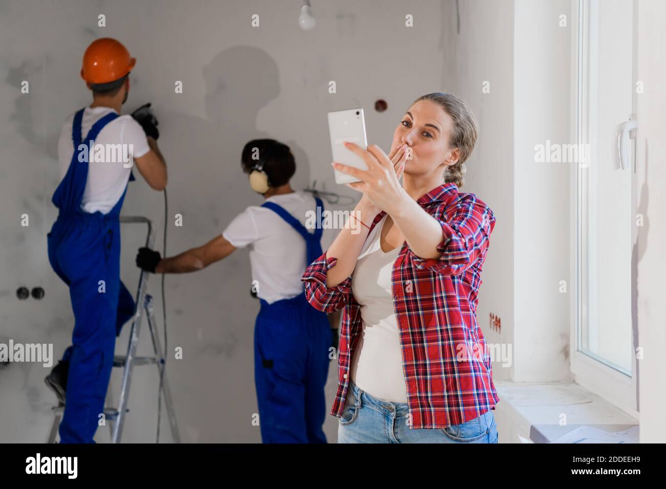 Une jeune femme sourit et prend un selfie au téléphone. En arrière-plan, un ouvrier général fore un mur Banque D'Images